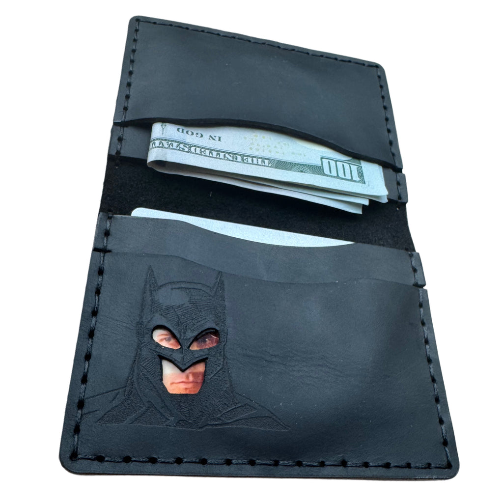 Open black leather bat bifold wallet with embossed superhero cowl and cutout eye window, holding cash and cards