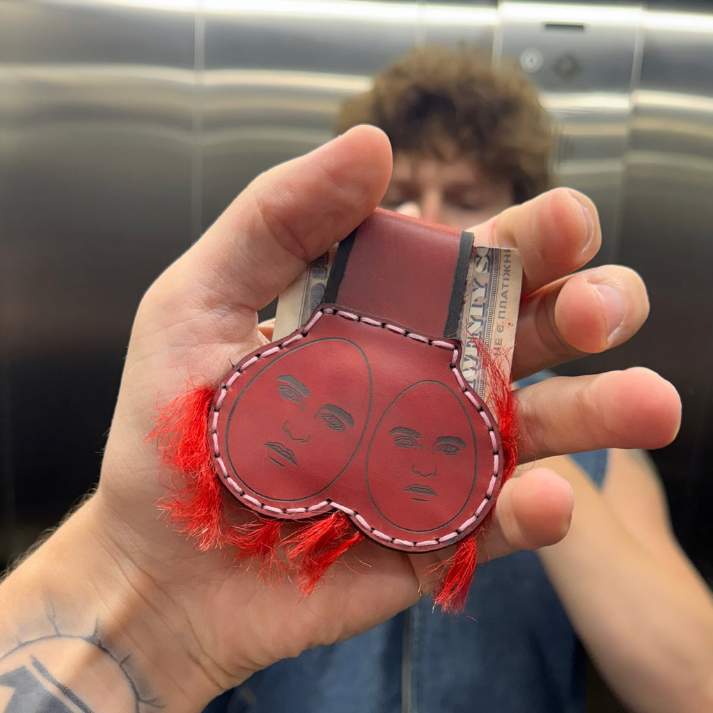 Red leather money clip with two hand-engraved faces and red fringe trim, held in a hand