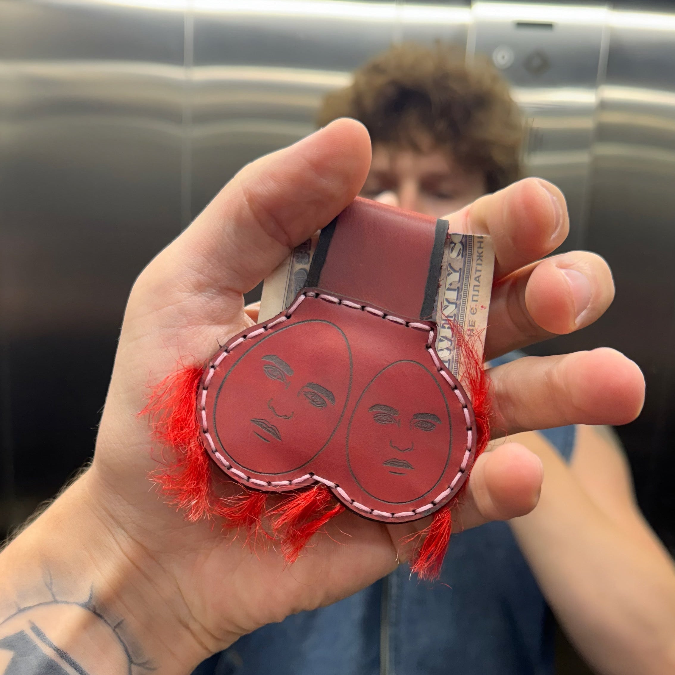 Red leather money clip with two hand-engraved faces and red fringe trim, held in a hand