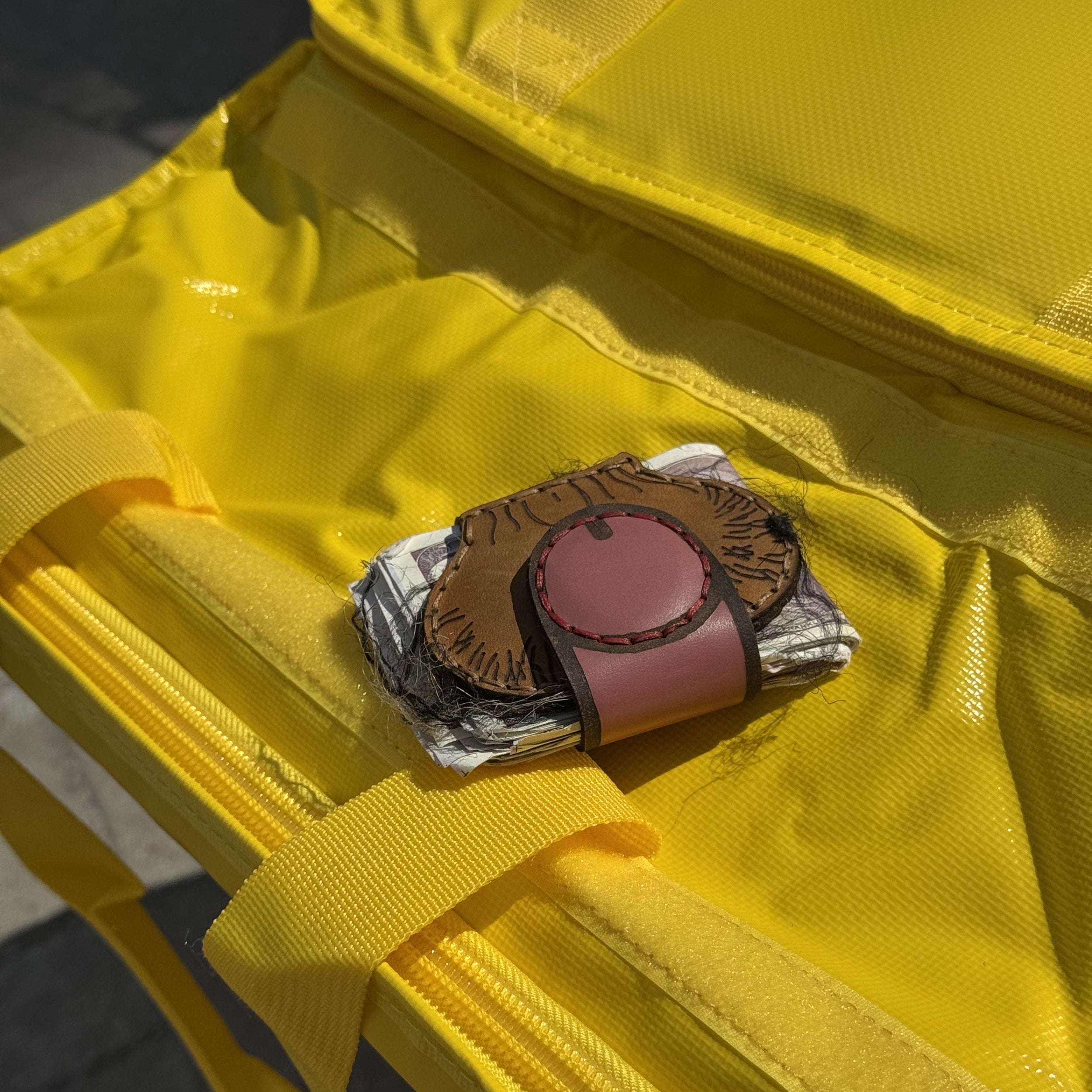 Brown leather XL cheeky money clip clamped on folded bills, sitting on a yellow bag